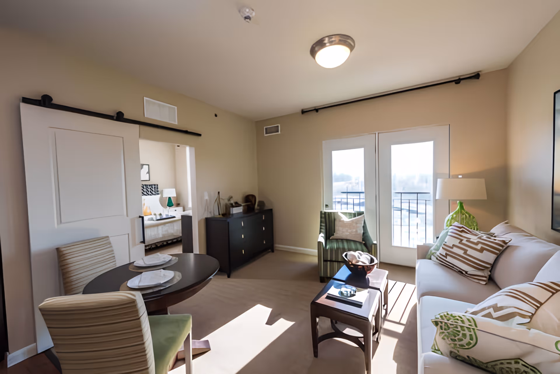 A bright living room with a white sofa adorned with patterned pillows, a green armchair, a small dark wood coffee table, and a round dining table with two striped chairs. There is a sliding barn door leading to a bedroom, and double glass doors letting in natural light.