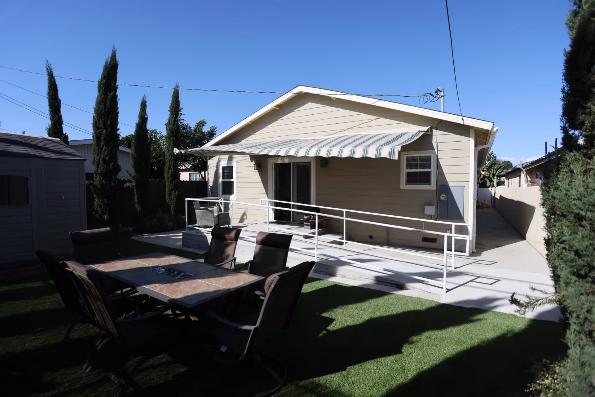 Outdoor patio area of a single-story building with beige siding, featuring a striped awning over a ramp with white railings. There is a table with six chairs on artificial grass in the foreground, surrounded by tall, narrow trees and a shed to the left.
