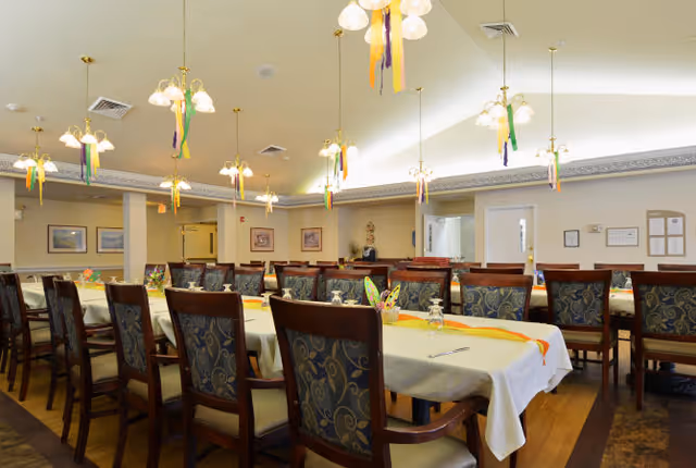 A spacious dining room with multiple tables covered in white tablecloths and decorated with colorful ribbons and small centerpieces. The room is well-lit with several hanging light fixtures and has wooden chairs with patterned upholstery arranged around the tables. The walls have framed pictures and there are doors and bulletin boards visible in the background.