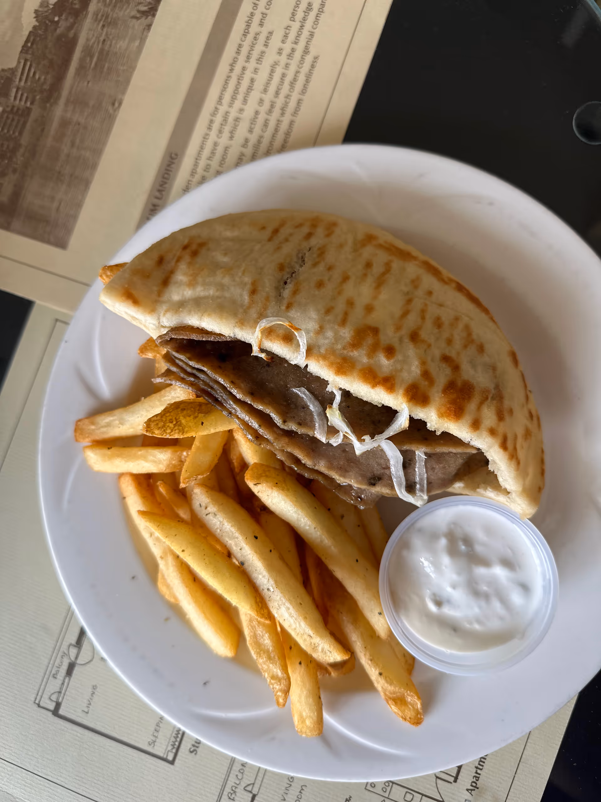 A white plate with a gyro sandwich containing sliced meat and onions, served with seasoned French fries and a small container of white dipping sauce, placed on a table with papers underneath.