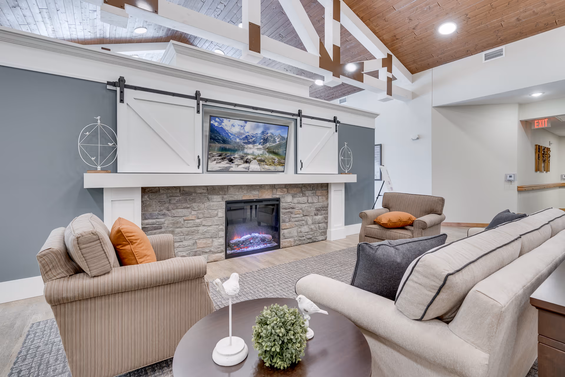 A cozy senior living common area with a stone fireplace, a mounted flat-screen TV above it, and comfortable seating including a beige sofa and two armchairs with orange and gray cushions. The room features a high wooden ceiling with exposed beams and soft lighting.