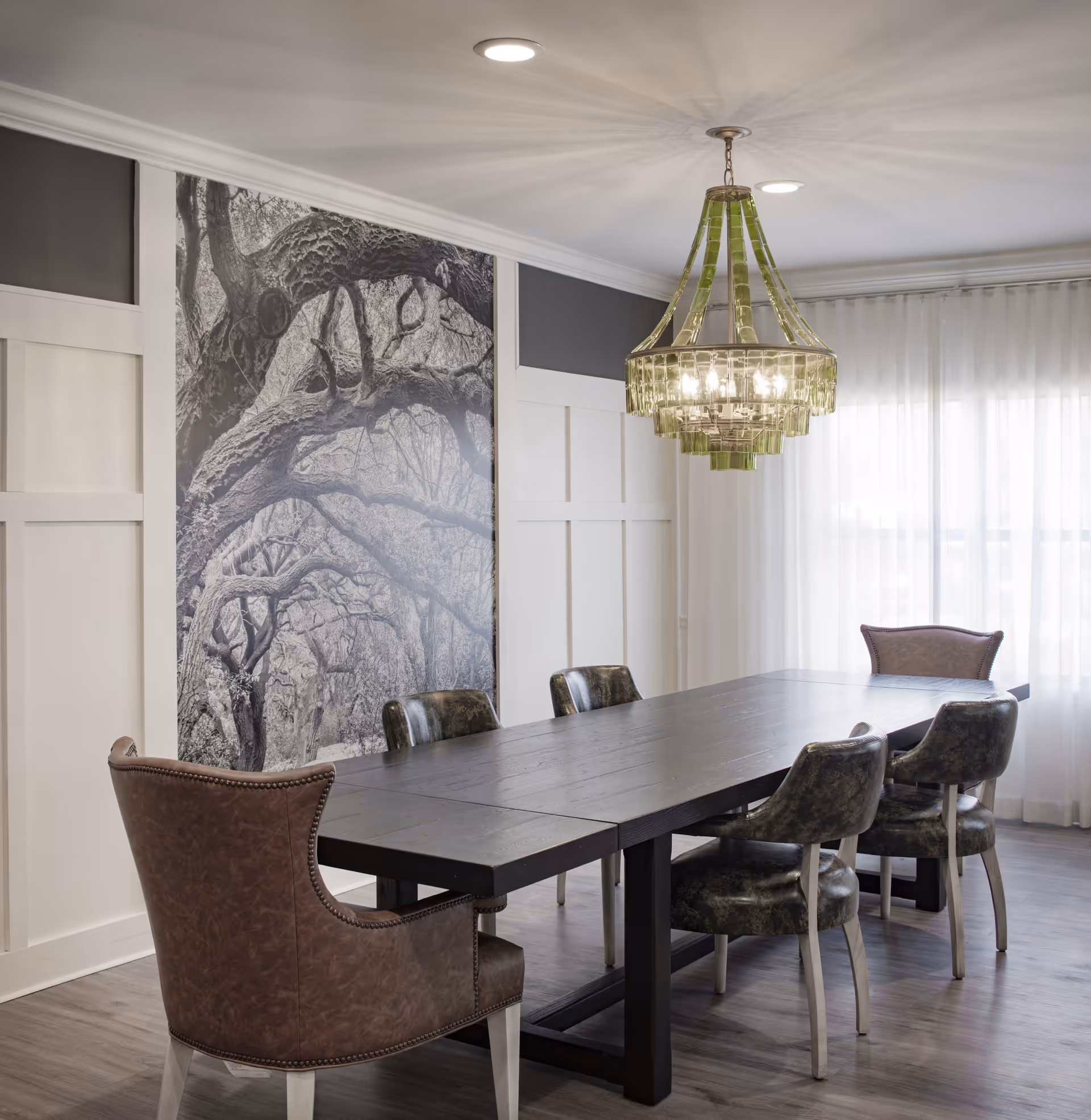 A modern dining room with a long dark wooden table surrounded by six chairs, including two brown leather armchairs and four black cushioned chairs. A large black and white photograph of tree branches decorates one wall, and a green chandelier hangs from the ceiling. Sheer white curtains cover the windows, allowing natural light to softly illuminate the room.