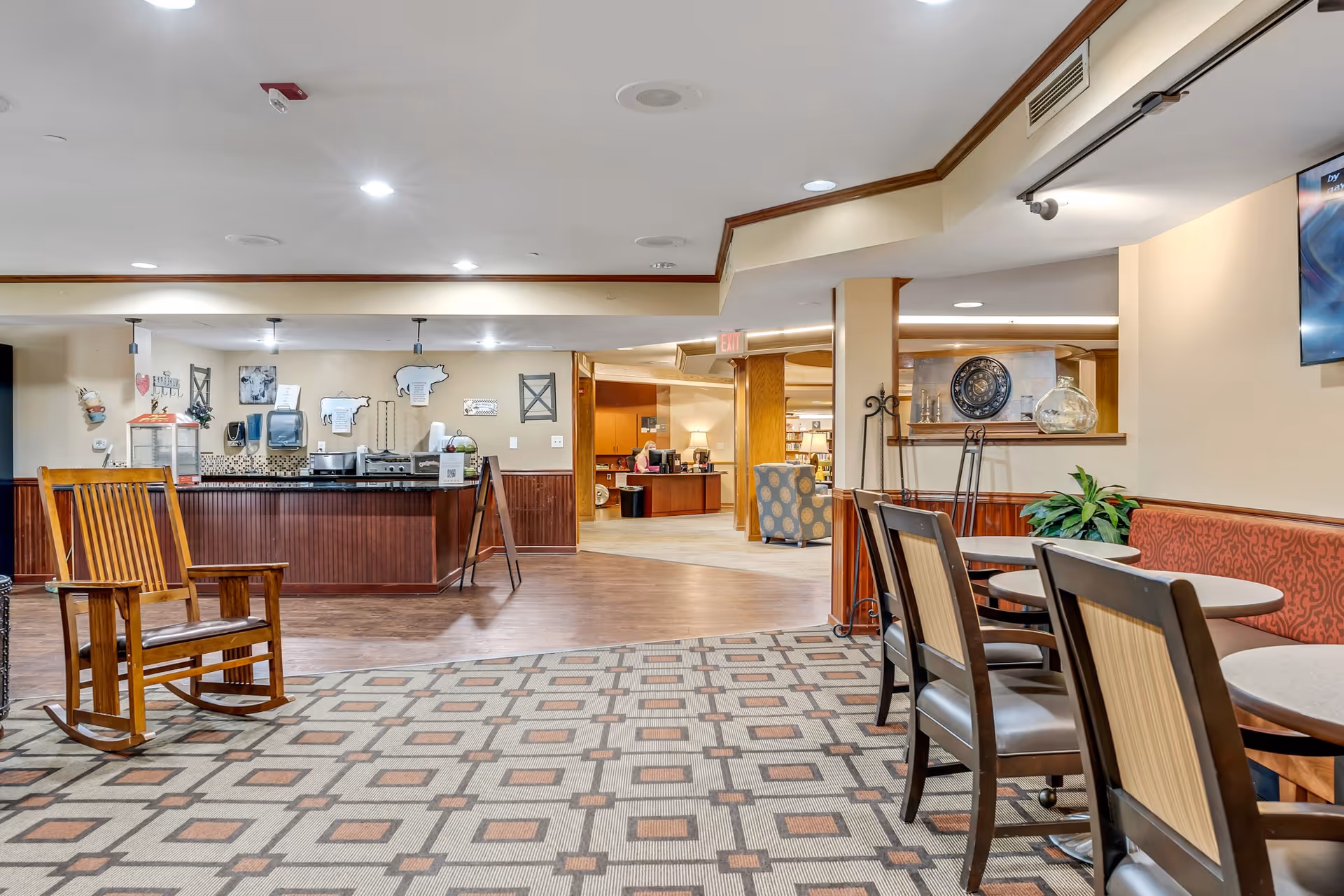 Interior view of a senior living facility common area with a patterned carpet, wooden rocking chair, tables and chairs along a wall with red patterned upholstery, a counter area with kitchen appliances and decorations on the wall, and a reception desk in the background.
