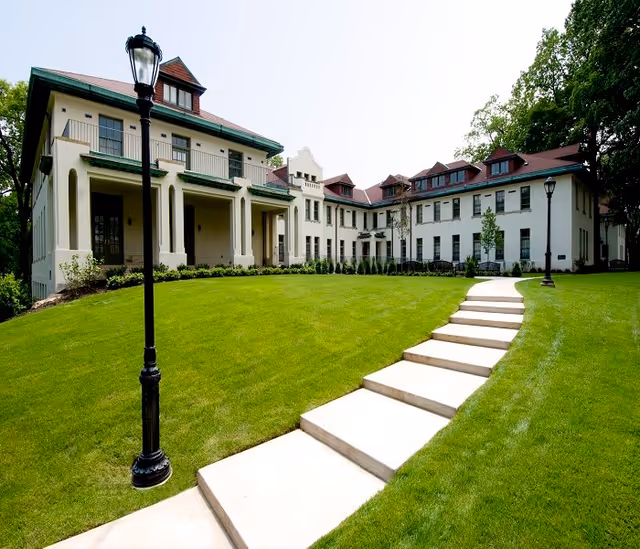 A large, elegant building with cream-colored walls and a red roof, surrounded by a well-maintained green lawn. A curved concrete pathway with steps leads up to the building entrance. There are two black street lamps along the pathway and trees in the background.