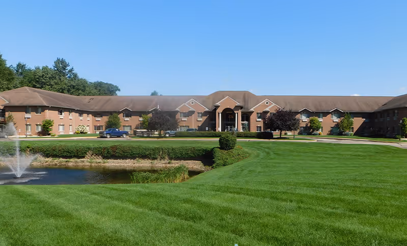 Wide exterior view of a two-story brick senior living facility building with a large green lawn, a small pond with a water fountain, and several trees and shrubs under a clear blue sky.