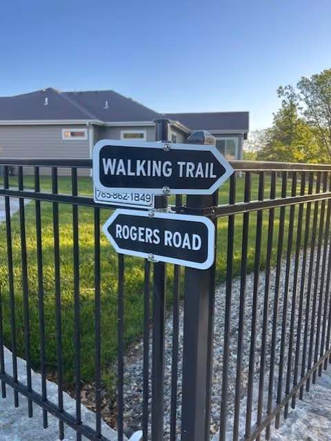 Black metal post with two directional signs attached, one pointing right labeled 'WALKING TRAIL' and the other pointing left labeled 'ROGERS ROAD', with a black metal fence and a house with a gray roof and beige siding in the background under a clear blue sky.