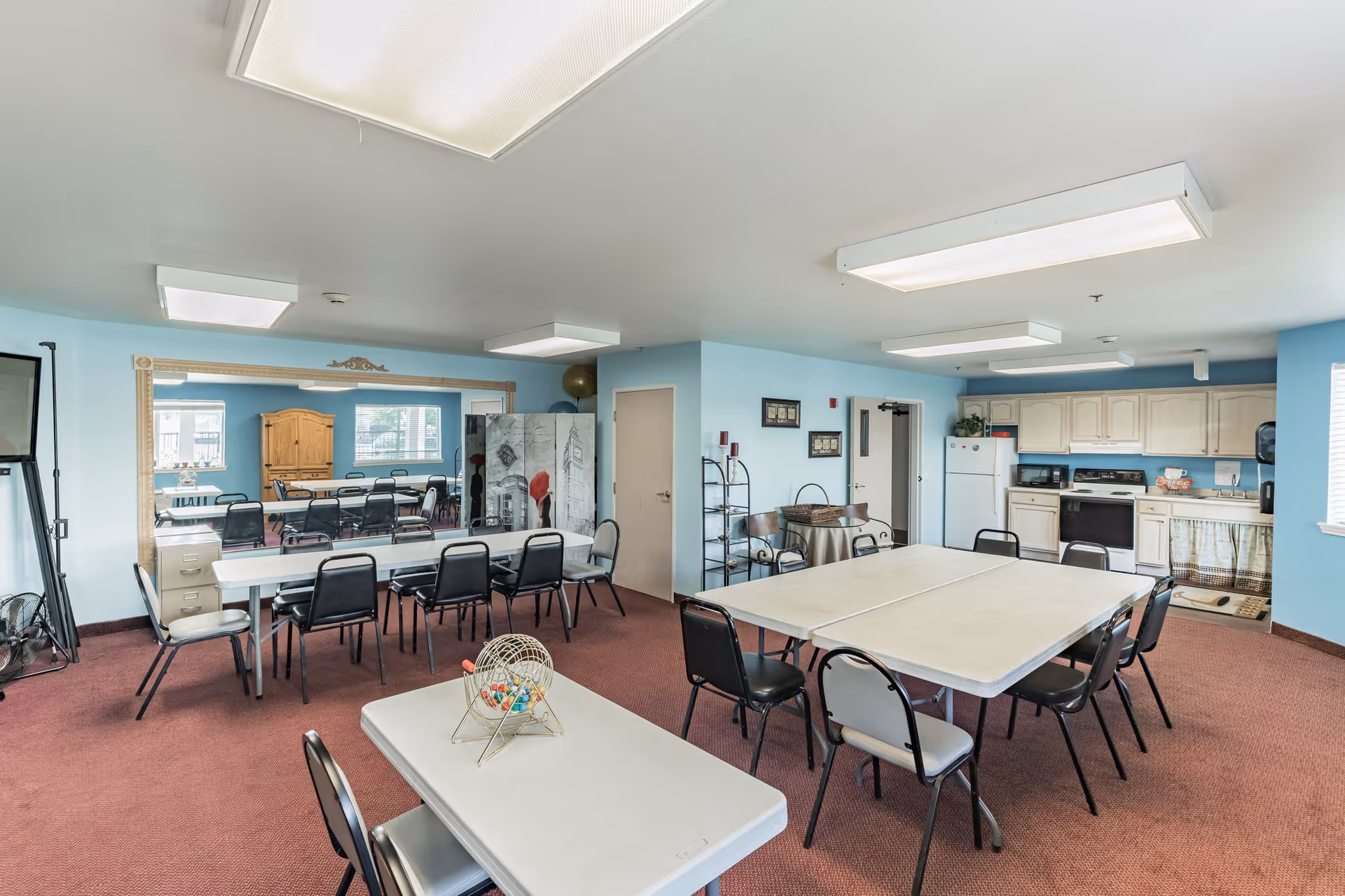 A spacious room with light blue walls and red carpet featuring several folding tables and chairs arranged for group activities. There is a kitchen area with white cabinets, a refrigerator, microwave, and stove in the background. A large mirror covers one wall, reflecting the room, and a bingo cage with colorful balls sits on one of the tables.