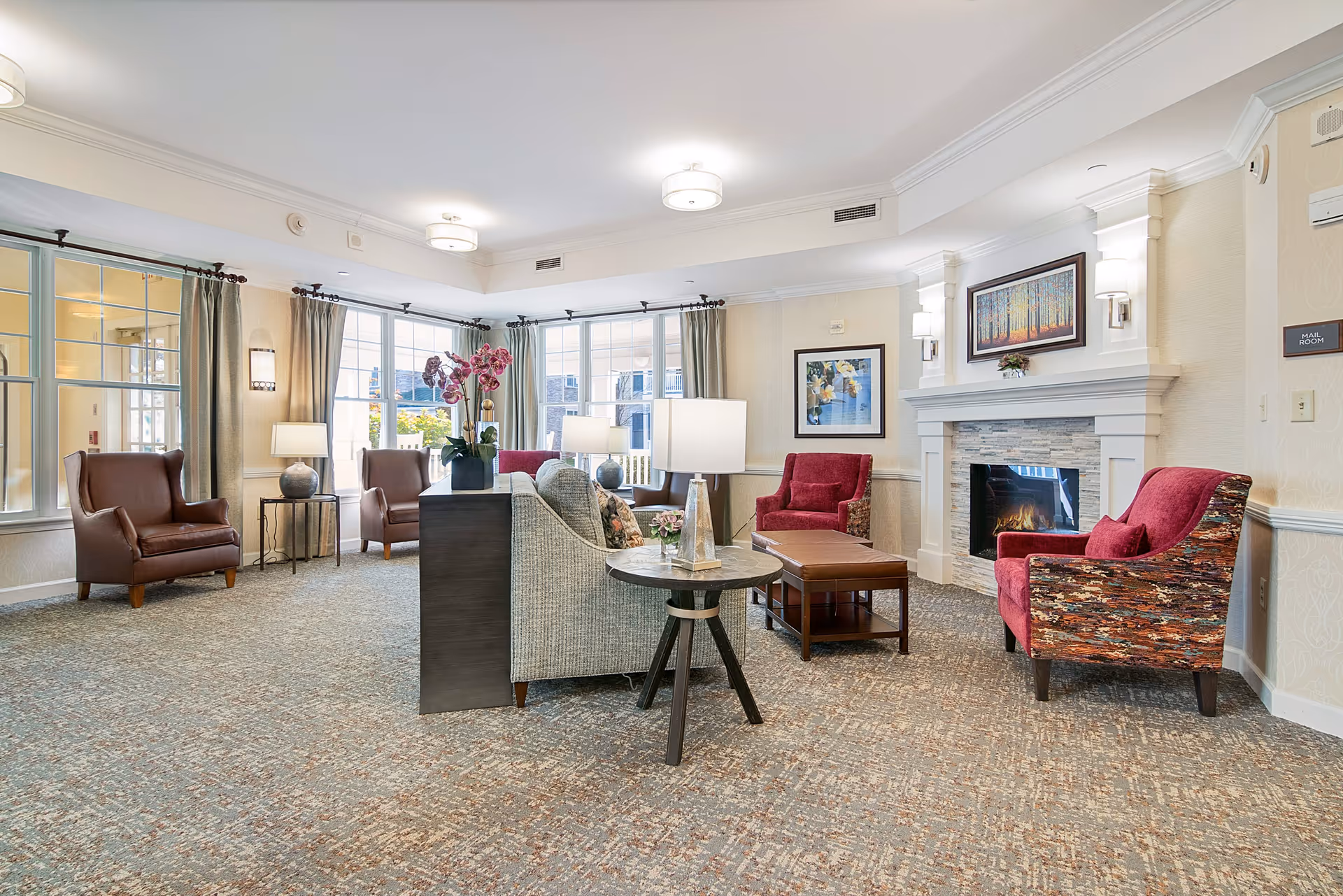 A bright and spacious living room area in a senior living facility with large windows, multiple armchairs in brown and red upholstery, a gray sofa, a fireplace with a painting above it, side tables with lamps, and a carpeted floor.