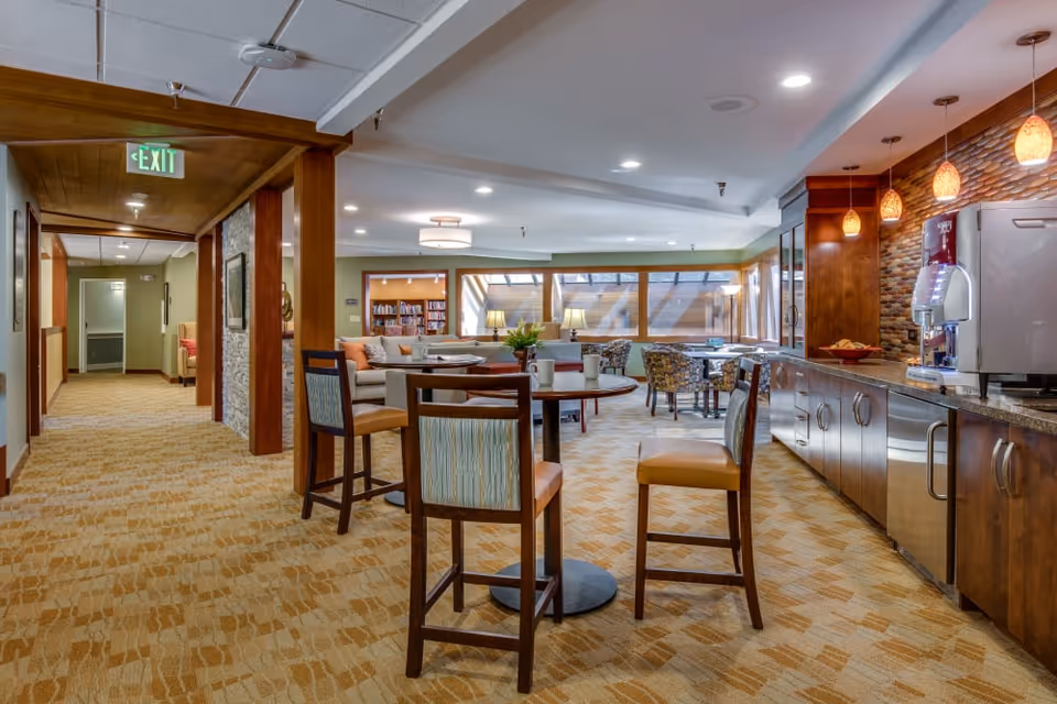 Open communal dining and lounge area with high-top tables and chairs, a kitchenette counter with pendant lights, and seating by large windows.