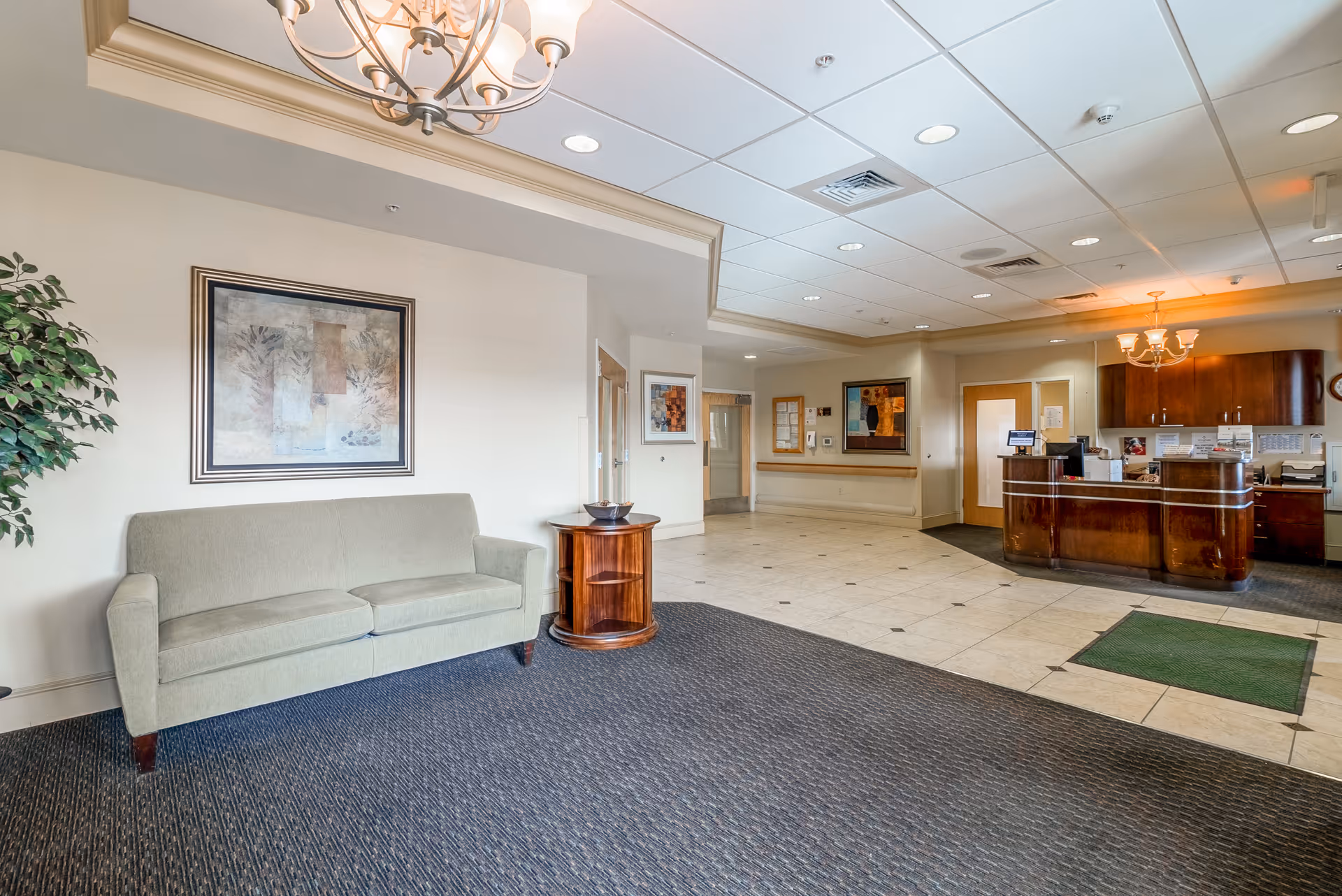 Interior view of a reception area in a senior living facility with a green sofa, a round wooden side table, framed artwork on the wall, a chandelier, and a wooden reception desk with cabinets behind it.