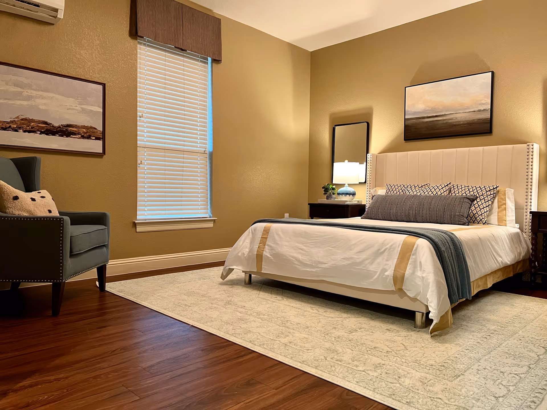 A neatly arranged bedroom with a bed featuring a white and beige bedspread, multiple pillows, and a gray throw blanket. There is a nightstand with a lamp and a small plant next to the bed. A large window with closed blinds is on the left wall, and a comfortable armchair with a pillow is positioned near the window. The walls are painted beige, and there are two framed artworks hanging on the walls. The floor is wooden with a large light-colored rug under the bed.