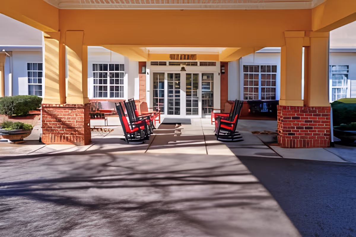 Covered entrance area of a building with yellow pillars and brick bases, featuring several black rocking chairs with red cushions arranged on either side of the walkway leading to white double doors with glass panes.