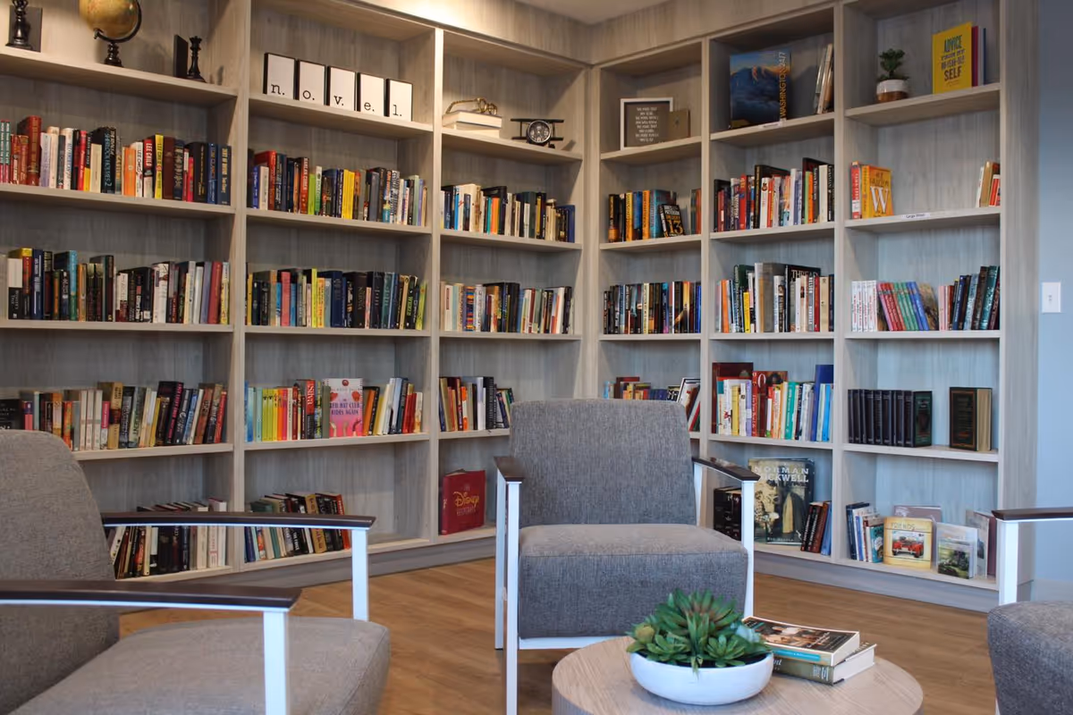 A cozy reading corner in a senior living facility featuring a large built-in bookshelf filled with books. There are three gray armchairs arranged around a small round wooden table with a potted succulent and a few books on it. The room has wooden flooring and a calm, inviting atmosphere.