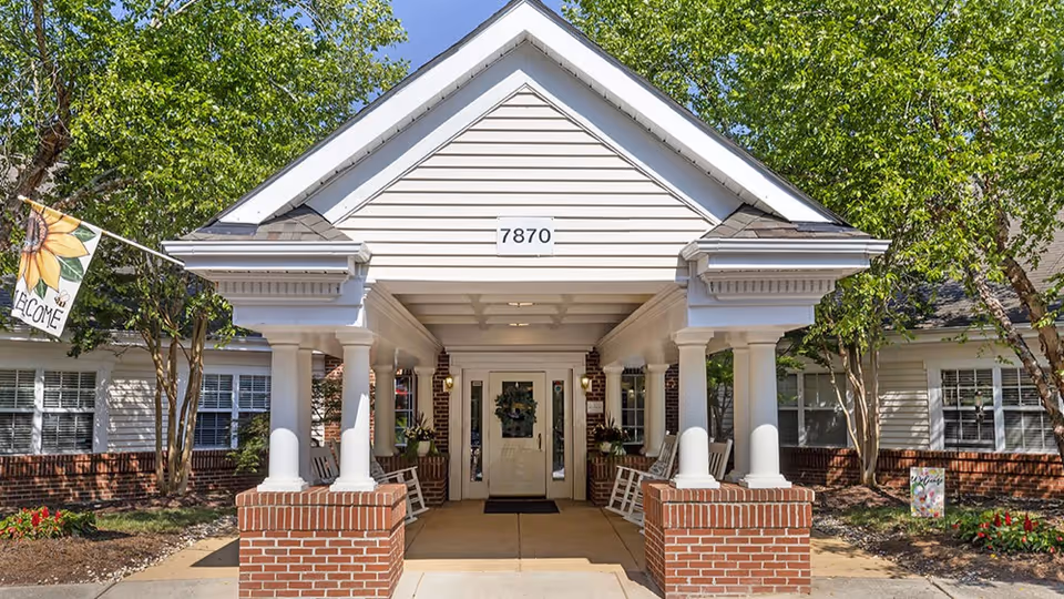 Entrance of a senior living facility with a covered porch supported by white columns on brick bases. The building has beige siding with the number 7870 displayed above the entrance door. There are rocking chairs on the porch and trees with green leaves on either side. A flag with a sunflower and the word 'Welcome' is visible on the left side.