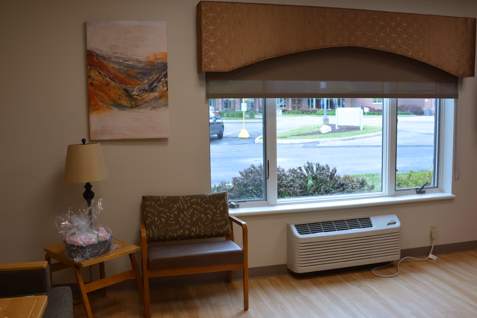 A small seating area inside a room with a wooden chair with patterned cushions, a wooden side table with a lamp and a wrapped gift basket, a large window with a beige valance showing a parking lot and greenery outside, and a white air conditioning unit below the window.