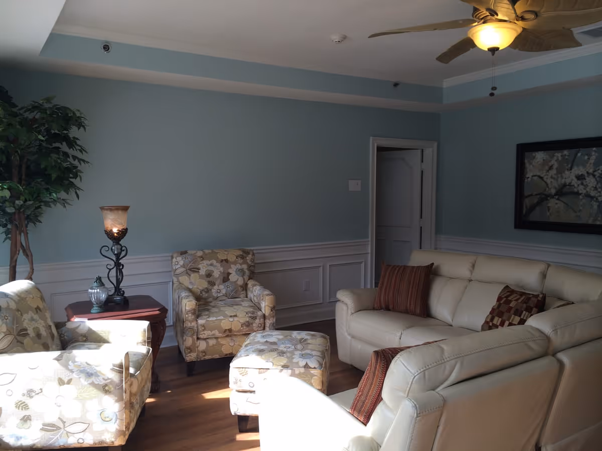 A cozy living room with light blue walls and white wainscoting. The room features a cream-colored leather sofa with two patterned throw pillows, two floral upholstered armchairs, and a matching ottoman. There is a wooden side table with a decorative lamp and a small glass ornament. A large potted plant is in the corner, and a framed floral painting hangs on the wall. The room has a ceiling fan with a light fixture and wooden flooring.