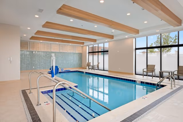Indoor community swimming pool with stainless handrails, seating, and large windows under wood-beamed ceiling.