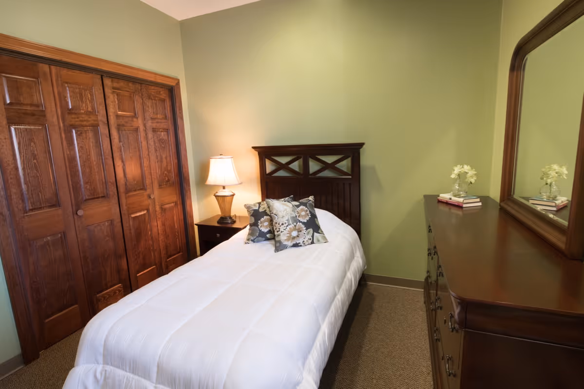 A small bedroom with a single bed covered in a white comforter and two floral pillows. Next to the bed is a wooden nightstand with a lamp. Across from the bed is a wooden dresser with a large mirror, decorated with a small vase of flowers and a couple of books. The walls are painted light green and there is a wooden closet with double doors.