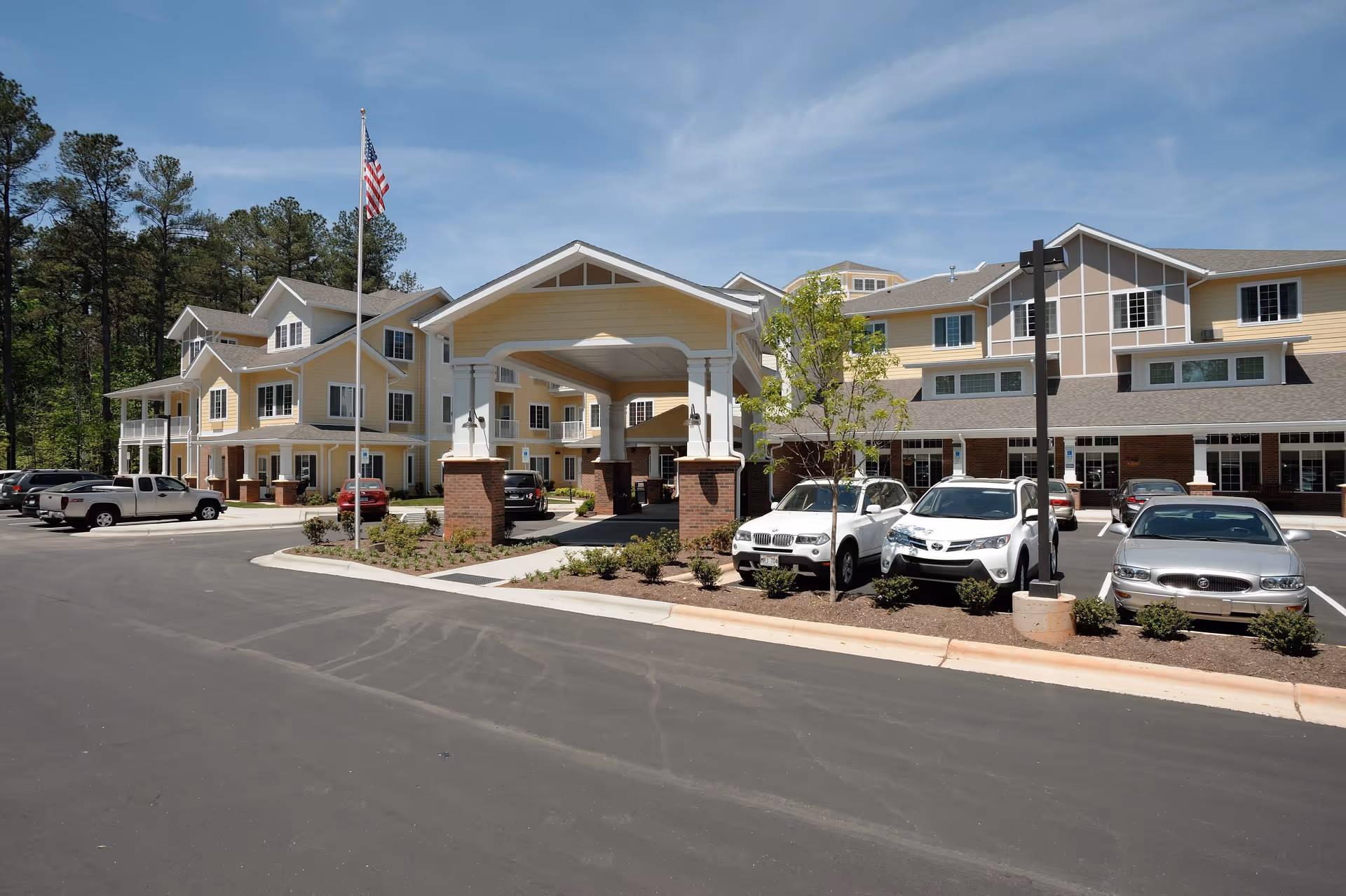 Exterior view of Whispering Pines Gracious Retirement Living facility showing a large multi-story building with a covered entrance, several parked cars, an American flag on a flagpole, and trees in the background under a clear blue sky.