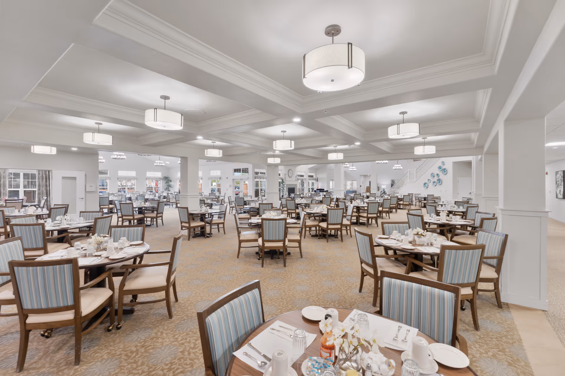 A spacious and well-lit dining room with multiple round tables set for meals. Each table has chairs with blue and beige striped upholstery, white tableware, and condiments. The room features a coffered ceiling with modern pendant lights and large windows allowing natural light to fill the space. The carpet has a subtle floral pattern, and there are decorative elements on the walls and a staircase visible in the background.