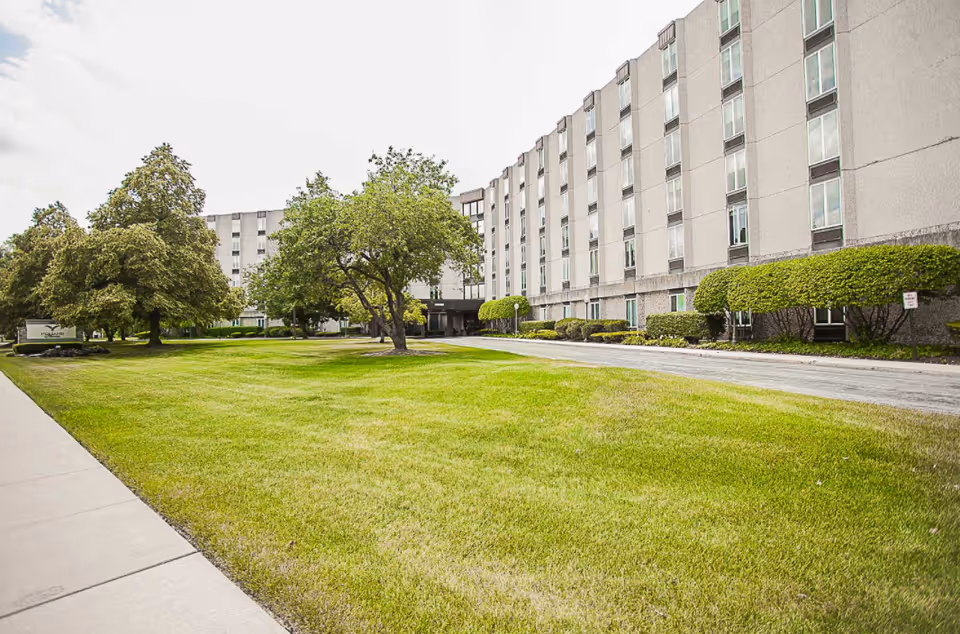 Exterior view of a multi-story senior living facility building with a well-maintained lawn, trees, and shrubs along a sidewalk and driveway under a partly cloudy sky.