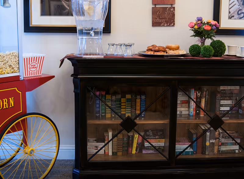 A cozy interior corner featuring a vintage-style popcorn machine with a popcorn container, a dark wooden cabinet with glass doors filled with books, a water dispenser with glasses on top, a tray with pastries, and a small flower arrangement and decorative greenery on the cabinet.