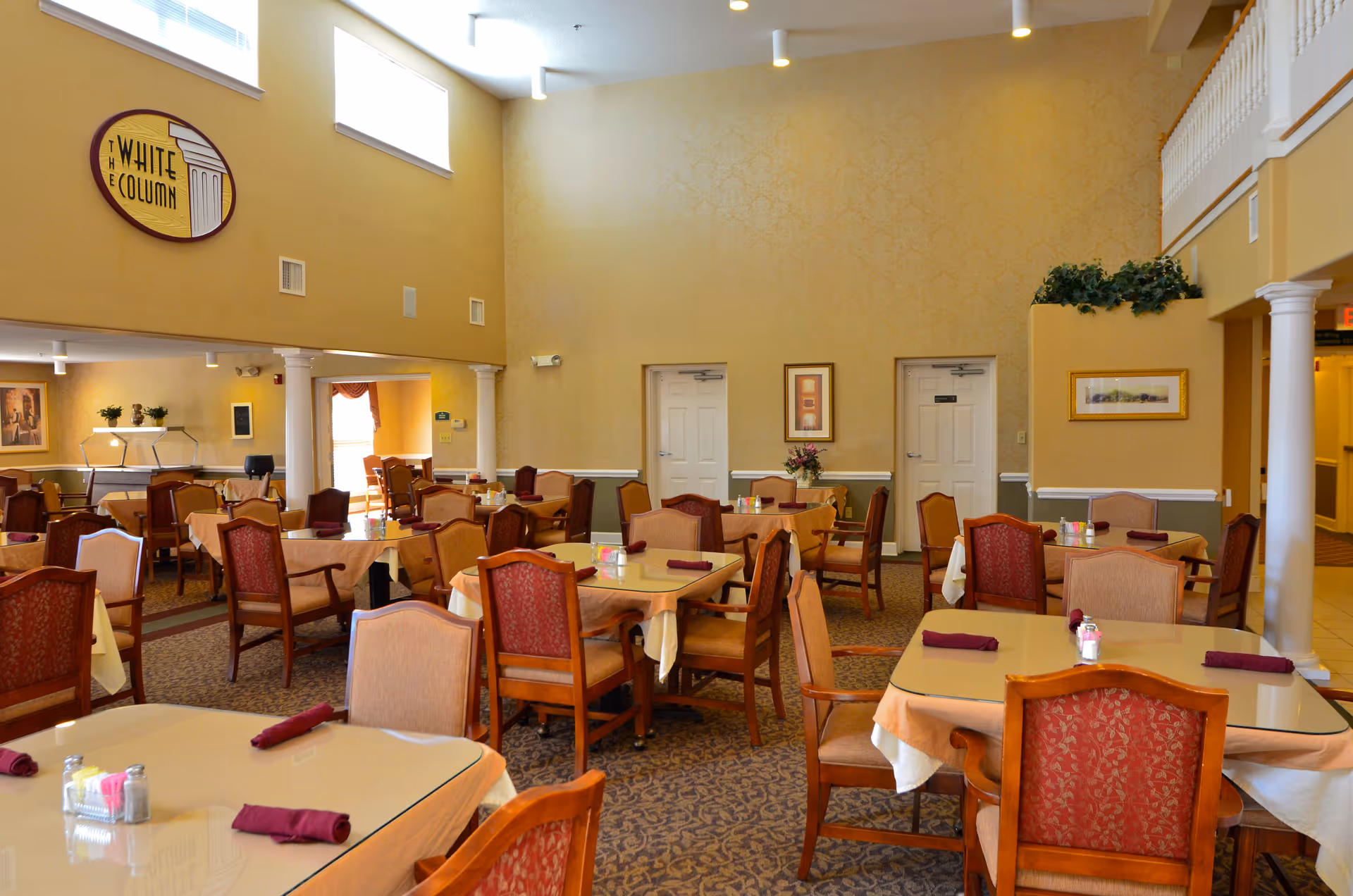 A spacious dining room with multiple tables covered with beige tablecloths and set with burgundy napkins. The room features high ceilings, yellow walls, decorative columns, framed artwork, and a sign that reads 'The White Column'. Chairs with red and beige upholstery surround the tables, and there is a carpeted floor with a patterned design.