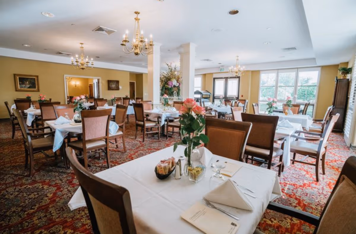 A bright and elegant dining room with multiple tables covered in white tablecloths, each set with napkins, glasses, and floral centerpieces. The room features chandeliers, large windows letting in natural light, and a patterned carpet.