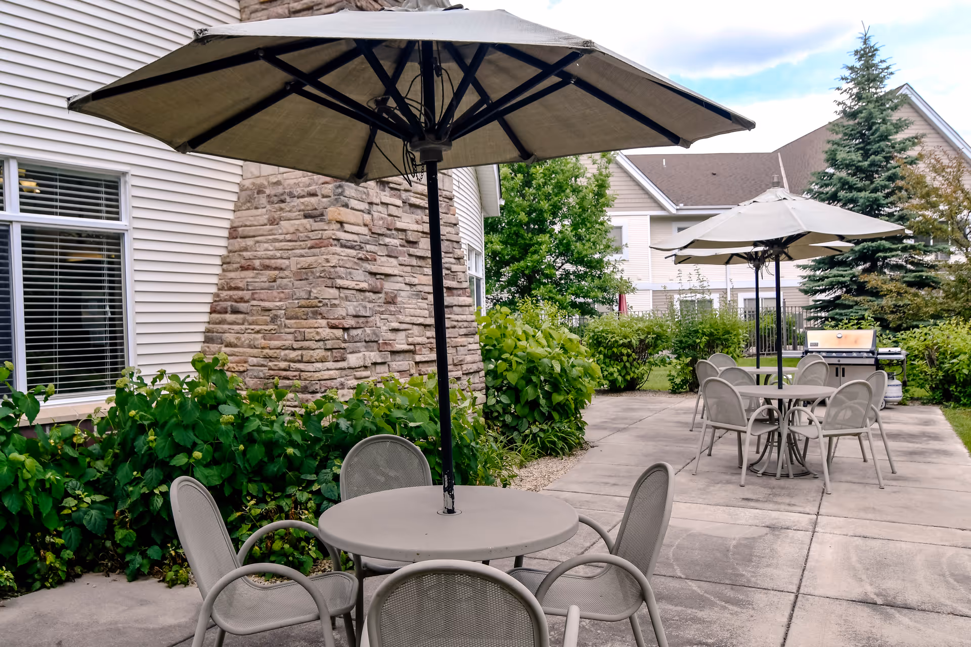 Outdoor patio with round tables, umbrellas, chairs, a grill, and surrounding landscaping beside a senior living building.
