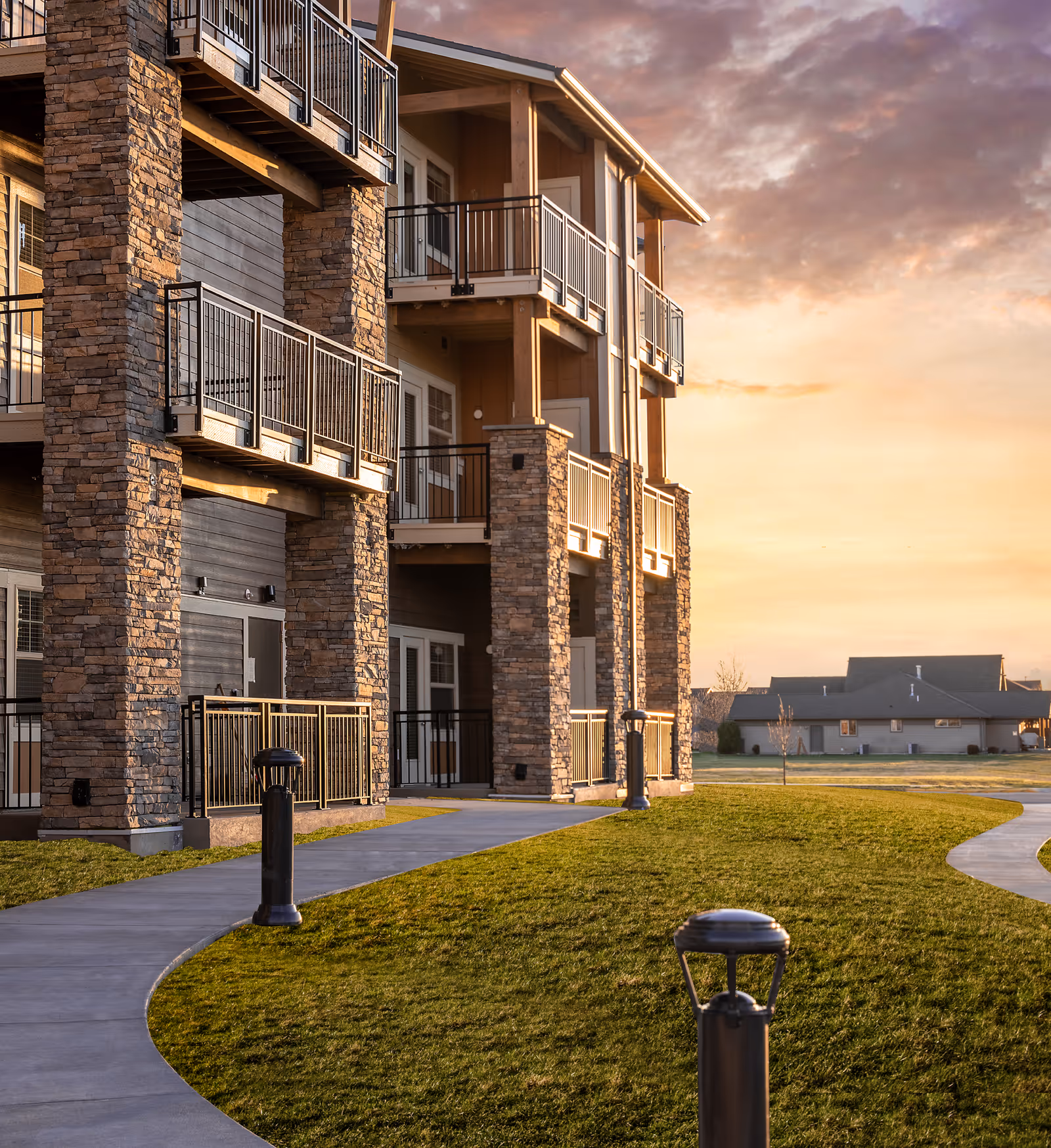Exterior view of a multi-story senior living facility building with stone pillars and balconies, a curved concrete walkway, green lawn, and a sunset sky in the background.
