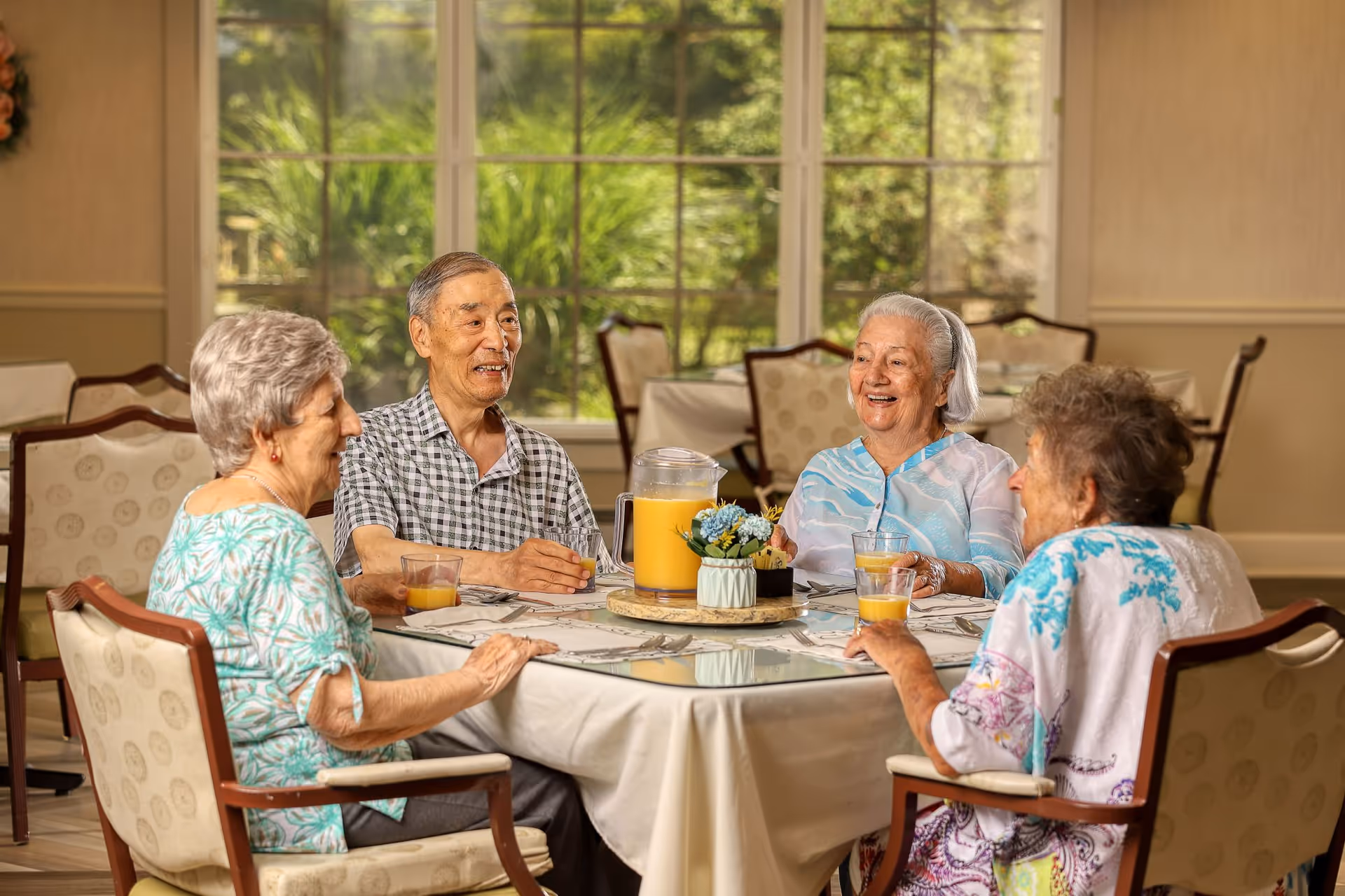 Four elderly people sitting around a dining table in a well-lit room with large windows showing greenery outside. They are smiling and holding glasses of orange juice. The table is covered with a white tablecloth and has a pitcher of orange juice and a small flower arrangement in the center.
