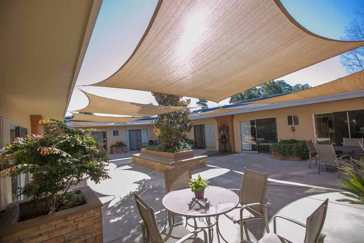 Outdoor courtyard area with shaded fabric canopies overhead, surrounded by single-story building walls with large windows and sliding doors. There are several patio tables and chairs, potted plants, and a raised planter with a tree in the center.