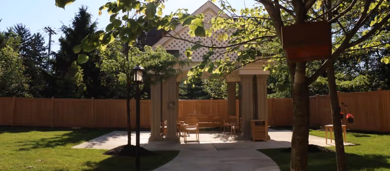 Outdoor seating area with a wooden pergola structure surrounded by a wooden fence and green grass. There are several chairs and tables under the pergola, with trees providing shade and a paved walkway leading to the seating area.