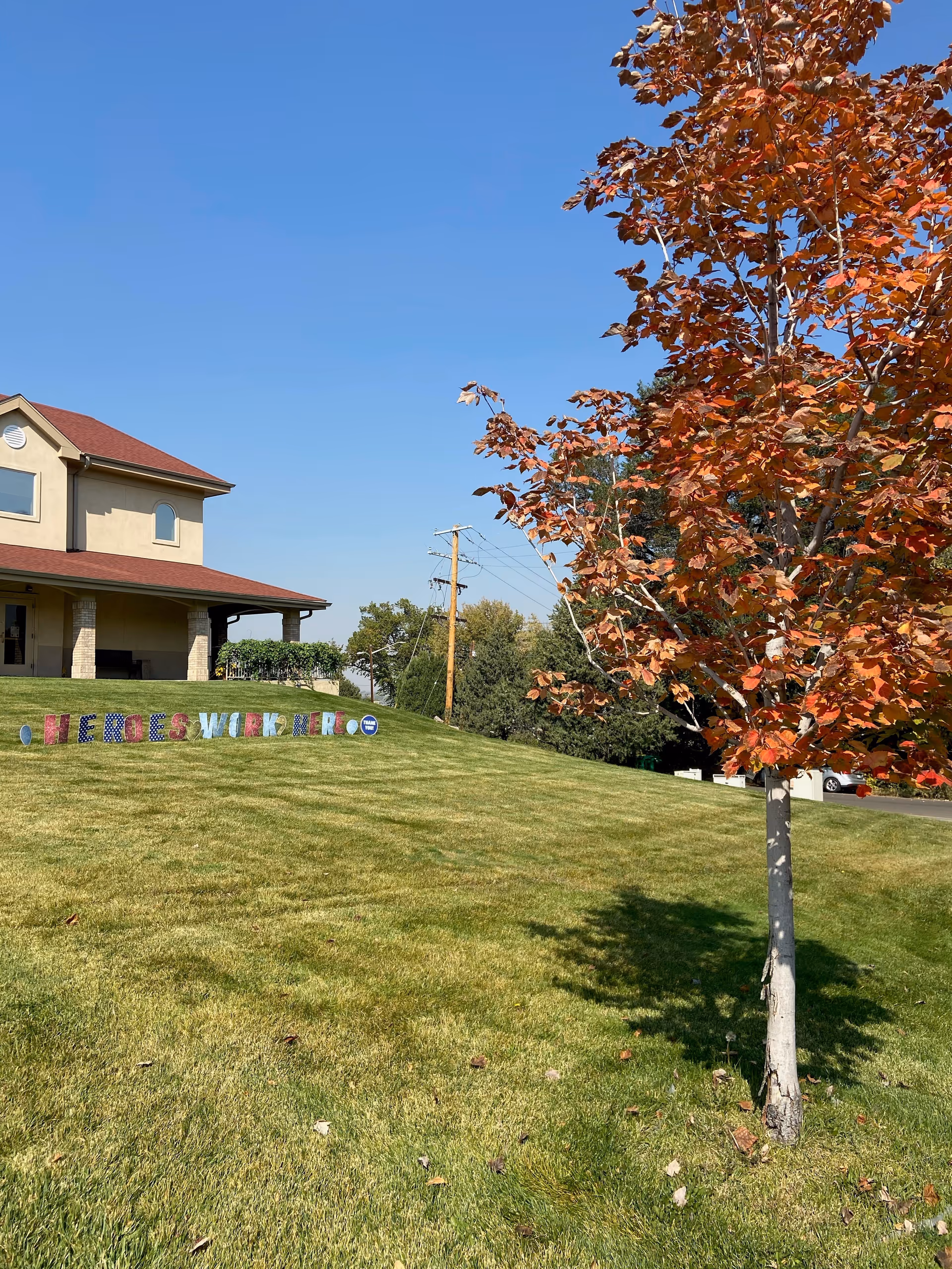 A grassy lawn with a tree showing autumn-colored leaves in the foreground and a beige two-story building with a red roof in the background under a clear blue sky. Colorful letters on the lawn spell out 'HEROES WORK HERE'.