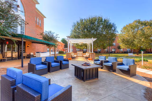 Outdoor patio area with blue cushioned wicker chairs arranged around a square fire pit. In the background, there is a white pergola with seating underneath and a brick building with green awnings. Trees and a well-maintained lawn surround the patio under a clear blue sky.
