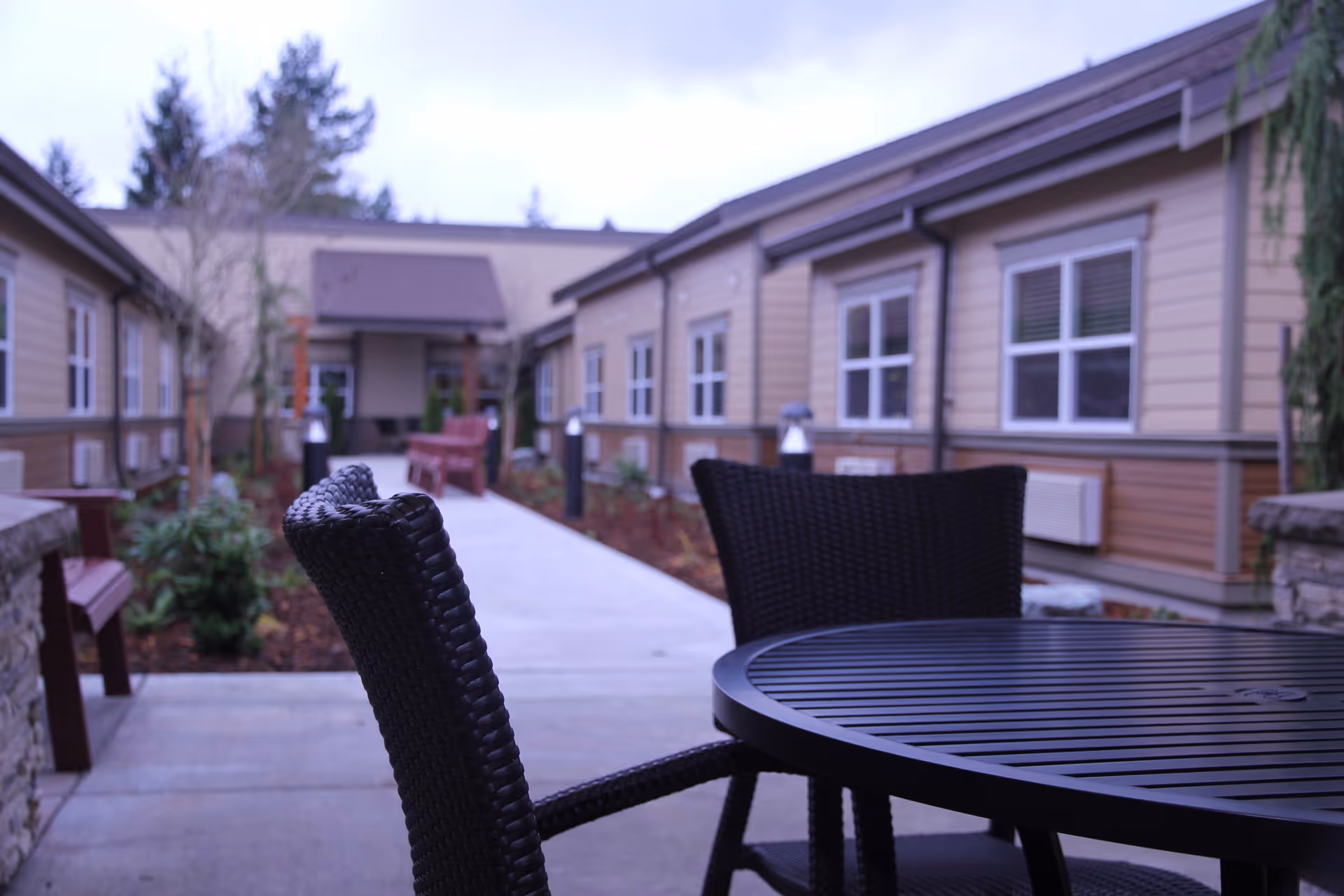 Outdoor courtyard area at Pine Ridge Post Acute with a round black metal table and two black wicker chairs in the foreground. The courtyard is surrounded by single-story buildings with beige siding and white-framed windows. There are benches, small trees, and plants along the concrete pathway leading to a covered entrance in the background.