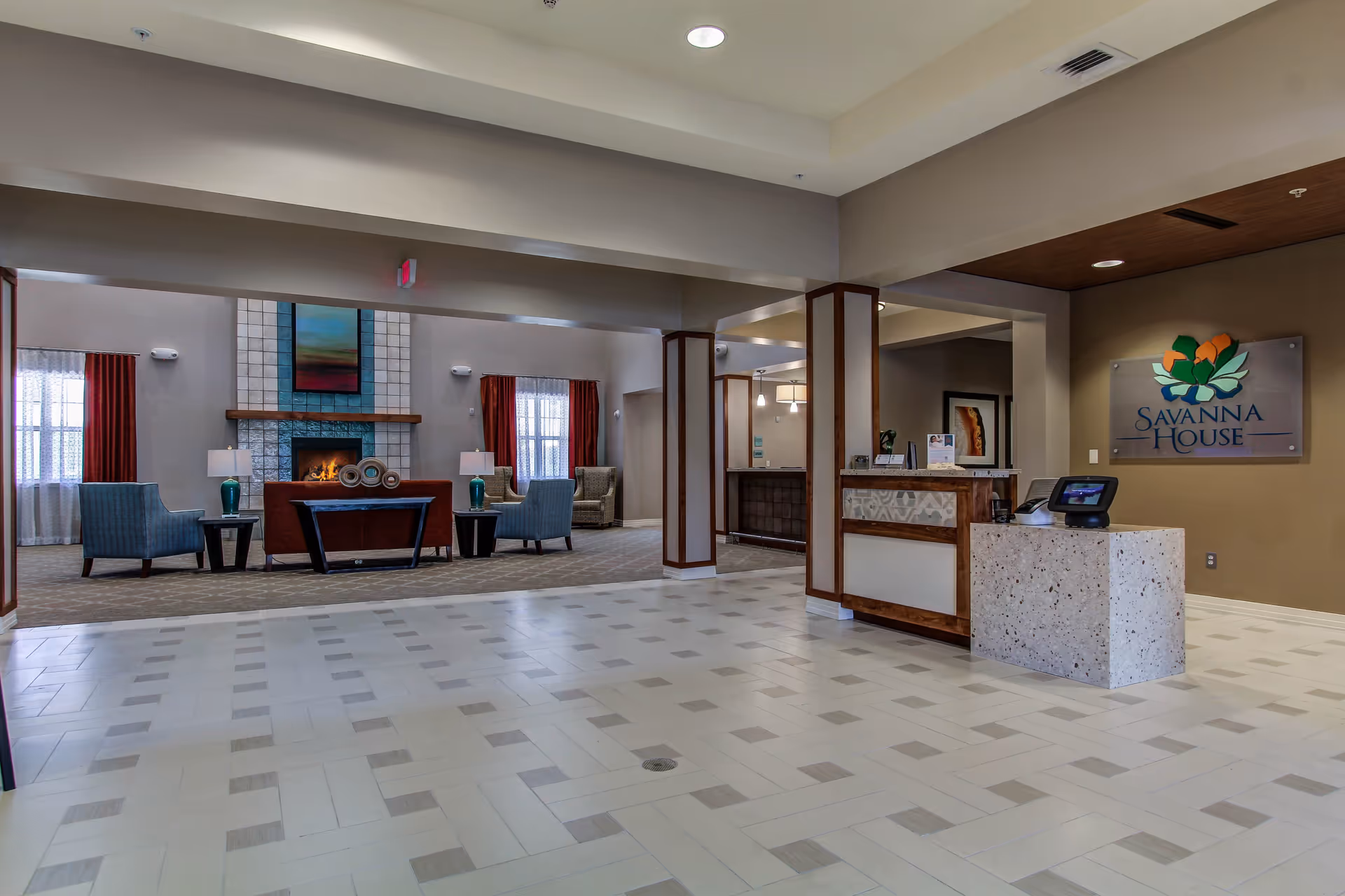 Interior view of Savanna House featuring a spacious lobby area with a reception desk on the right and a cozy seating area with chairs, a sofa, lamps, and a fireplace in the background. The floor has a light-colored tile pattern and the walls are painted in neutral tones.