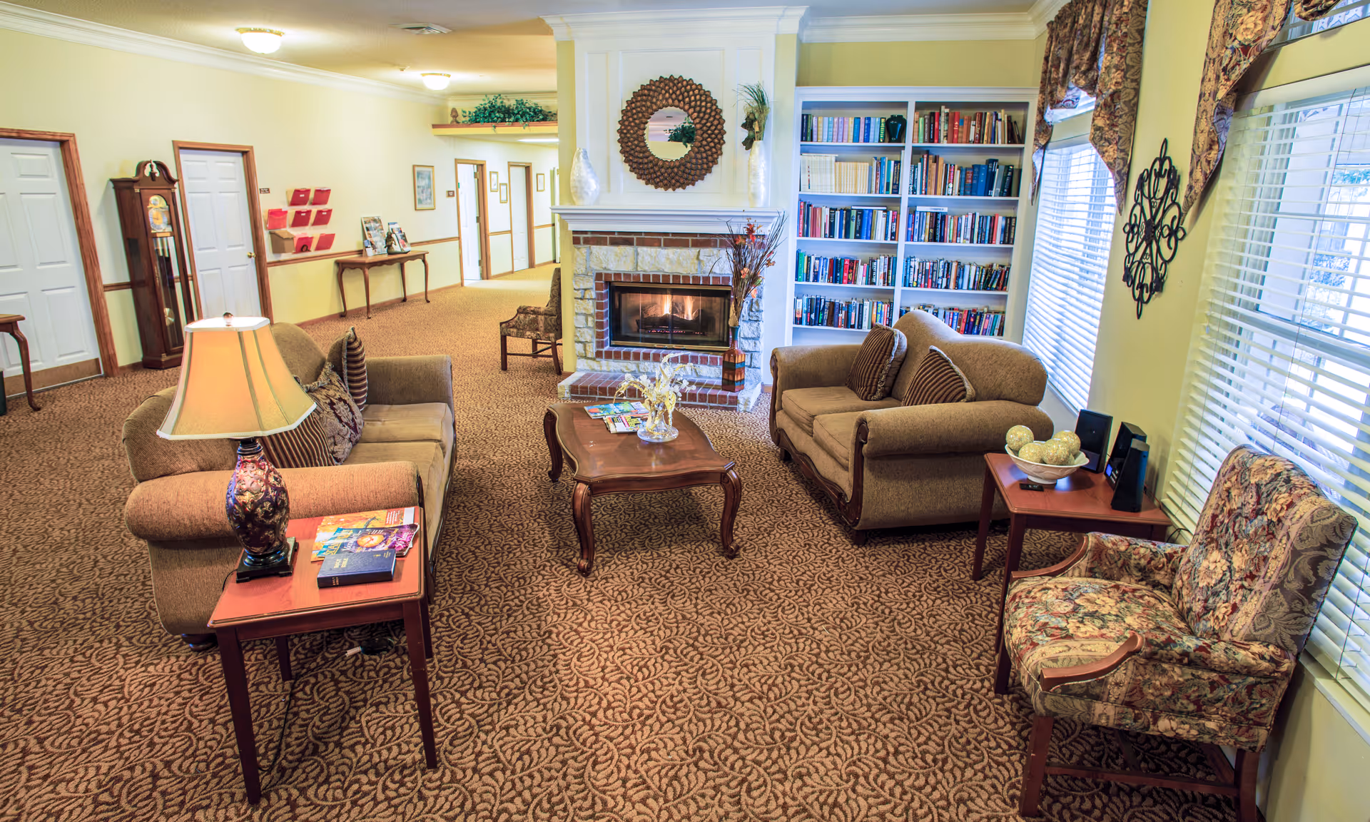 A cozy living room area in a senior living facility with two brown sofas, a floral armchair, a wooden coffee table, and side tables with decorative items. There is a lit fireplace with a round decorative mirror above it and a built-in bookshelf filled with books. Large windows with patterned curtains allow natural light to enter the room. The carpet has a leaf pattern, and the walls are painted light yellow. A hallway with doors and a grandfather clock is visible in the background.