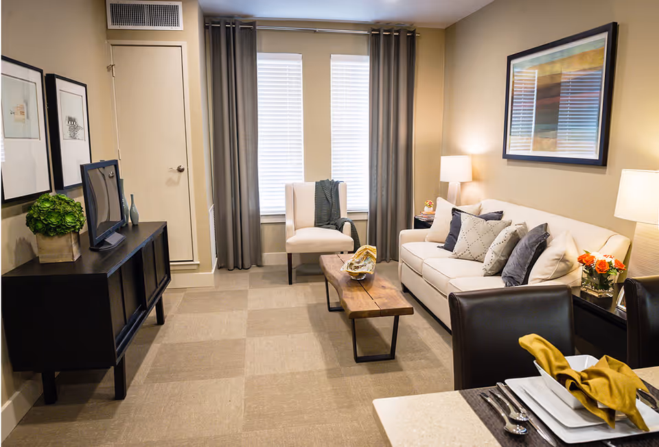 Cozy furnished living room with a sofa, armchair, coffee table, TV console, and a dining place visible in the foreground.