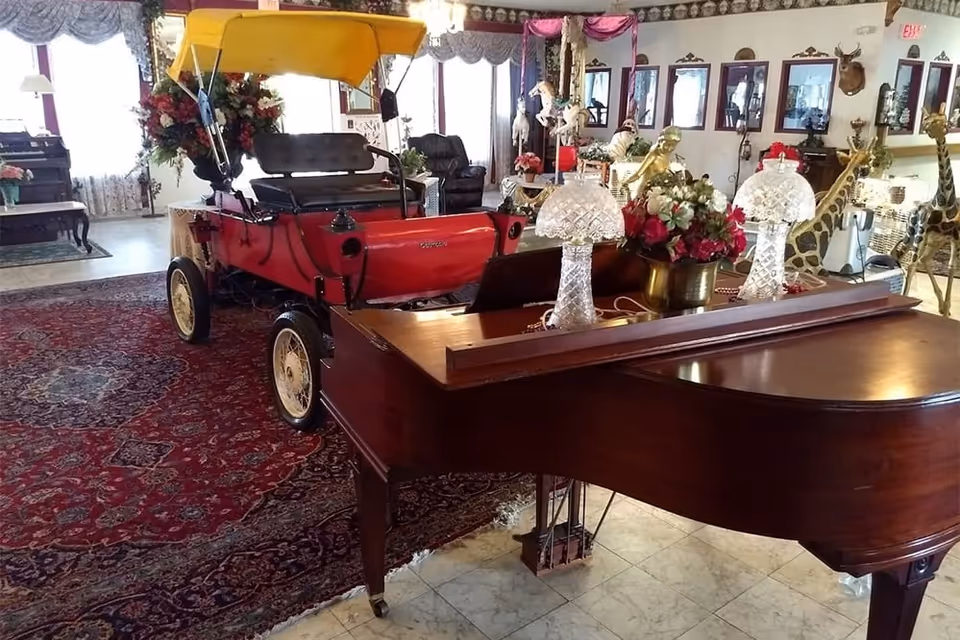 Interior room with a vintage red car with a yellow canopy displayed on a large red patterned rug. In the foreground is a wooden grand piano with two crystal lamps and a flower arrangement on top. The room is decorated with various floral arrangements, animal figurines including giraffes, and framed pictures on the walls.