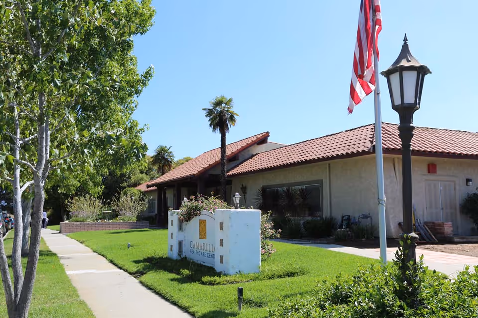 Exterior view of Camarillo Healthcare Center building with a tiled roof, a white sign with the facility name, an American flag on a flagpole, a vintage-style street lamp, green grass, trees, and a clear blue sky.
