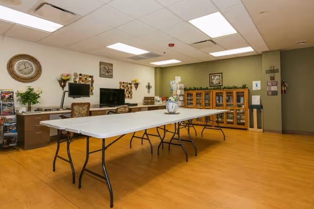 A spacious room with wooden flooring and a long white folding table in the center. The walls are decorated with a large clock, framed pictures, and decorative wall hangings. There is a cabinet with glass doors filled with items, a computer monitor on a desk, and chairs placed around the room. The ceiling has fluorescent lighting panels.