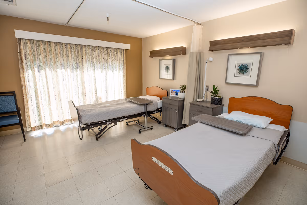 Two adjustable healthcare beds in a bright shared patient room with nightstands, chairs, and a large curtained window.