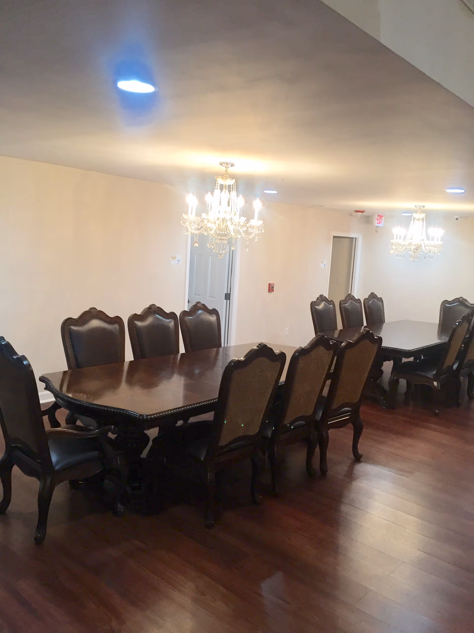A dining room with two long dark wooden tables surrounded by matching high-back chairs with cushioned seats and backs. The room has wooden flooring, beige walls, and two ornate chandeliers hanging from the ceiling providing warm lighting.