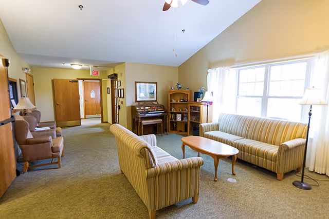 A bright communal living room with striped sofas, armchairs, a coffee table, a piano and a large window.
