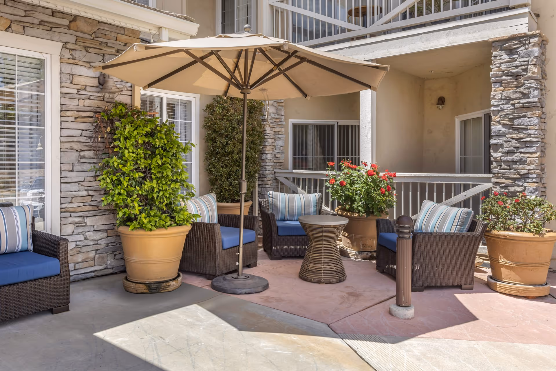 Outdoor patio area with four wicker chairs featuring blue cushions and striped pillows arranged around a small round wicker table. A large beige umbrella provides shade. Several large potted plants with green foliage and red flowers decorate the space. The patio is adjacent to a building with stone and stucco walls and sliding glass doors.