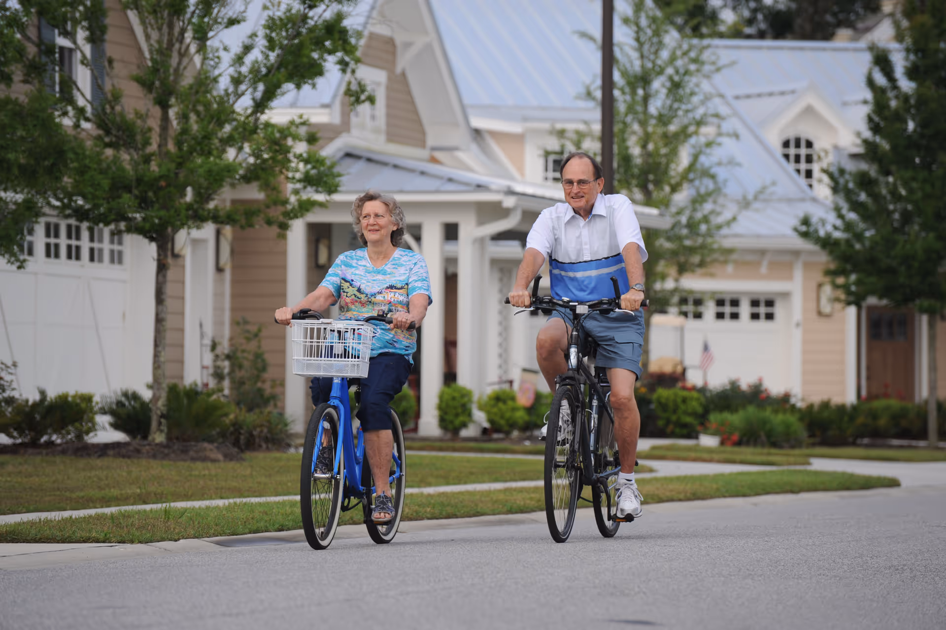 An elderly woman and man riding bicycles on a residential street in a neighborhood with houses and greenery in the background.