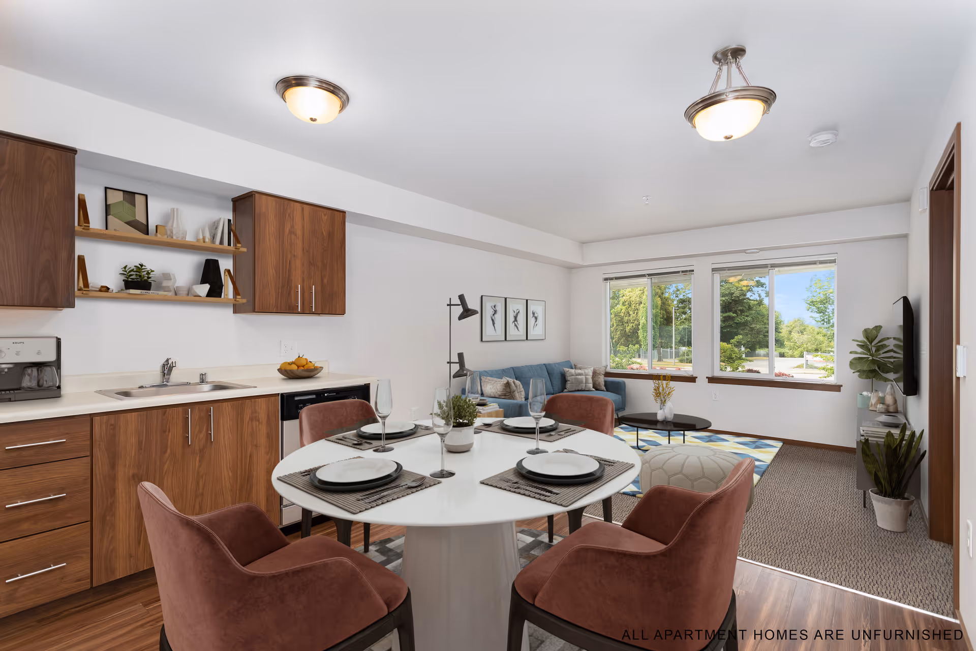 Interior view of a senior living apartment at Reserve at SeaTac featuring a small kitchen area with wooden cabinets, a white round dining table set for four with pink upholstered chairs, and a living room area with a blue sofa, coffee table, floor lamp, and large windows showing greenery outside.