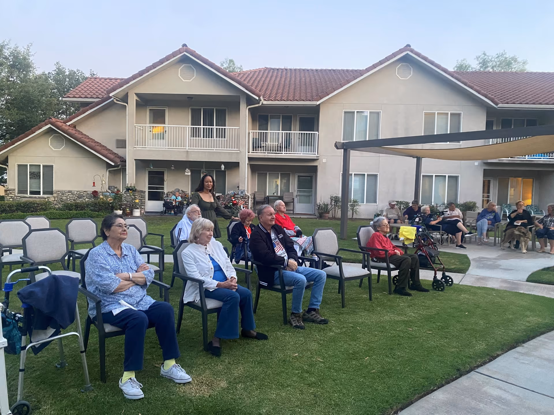 A group of elderly people sitting on chairs outdoors on a lawn in front of a two-story residential building with balconies. Some individuals are using walkers, and a woman is standing among them. The setting appears to be a social or recreational gathering at a senior living facility.