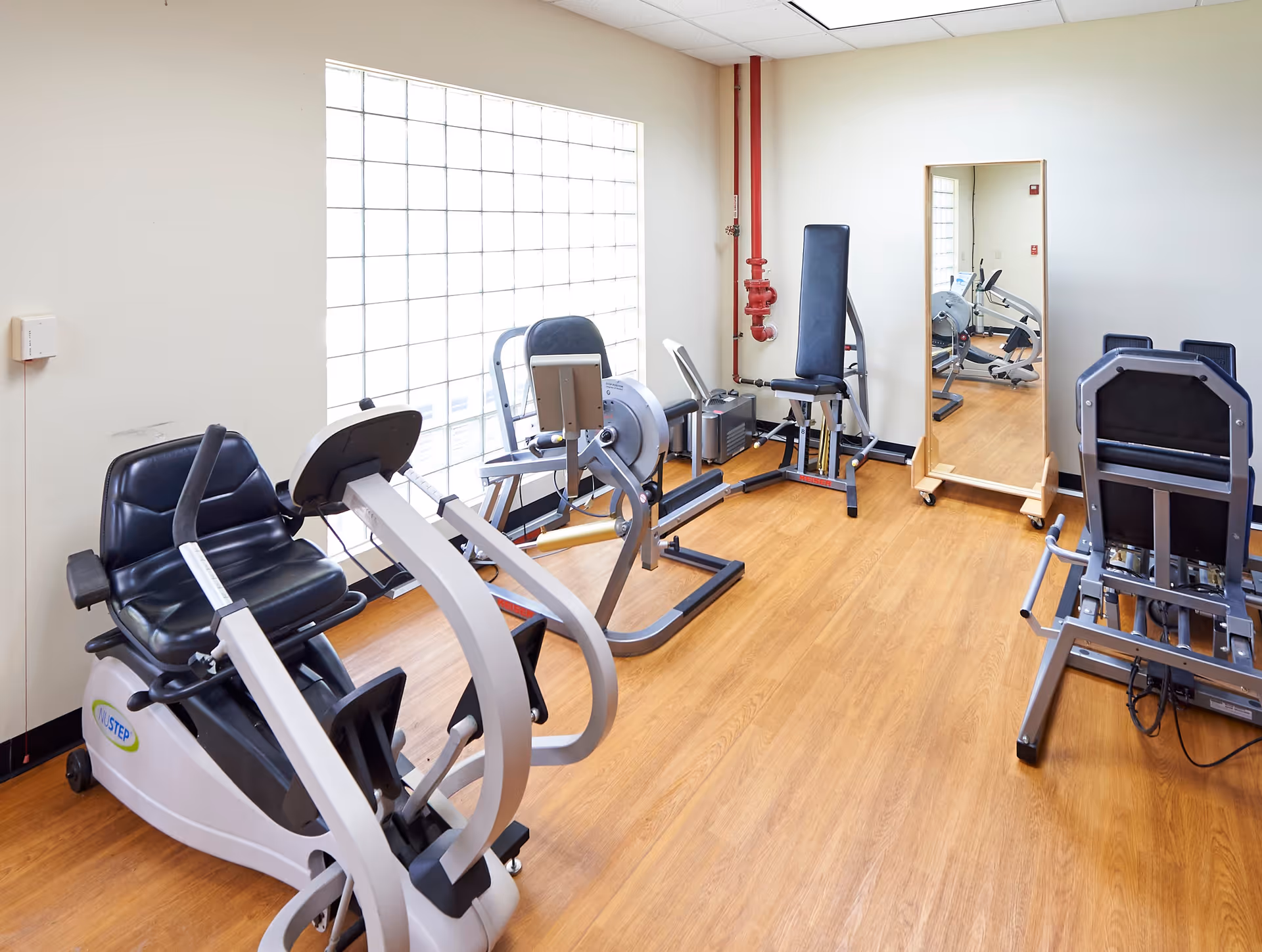 A small fitness room with exercise equipment including a recumbent bike, a rowing machine, and a leg press machine. The room has a large glass block window allowing natural light, a wooden floor, a full-length mirror on wheels, and white walls with a red fire sprinkler pipe.
